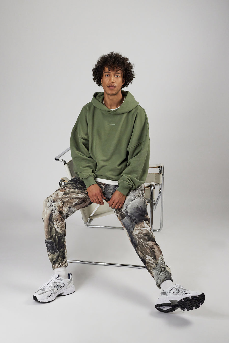 Man sitting on a chair wearing allover printed sweatpants and hoddie in color sage green
