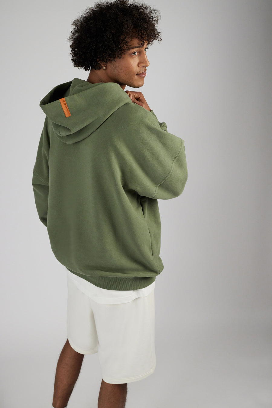 Man wearing shorts in color eggnog and a hoodie in color sage green with a small labeling detail at backside hood